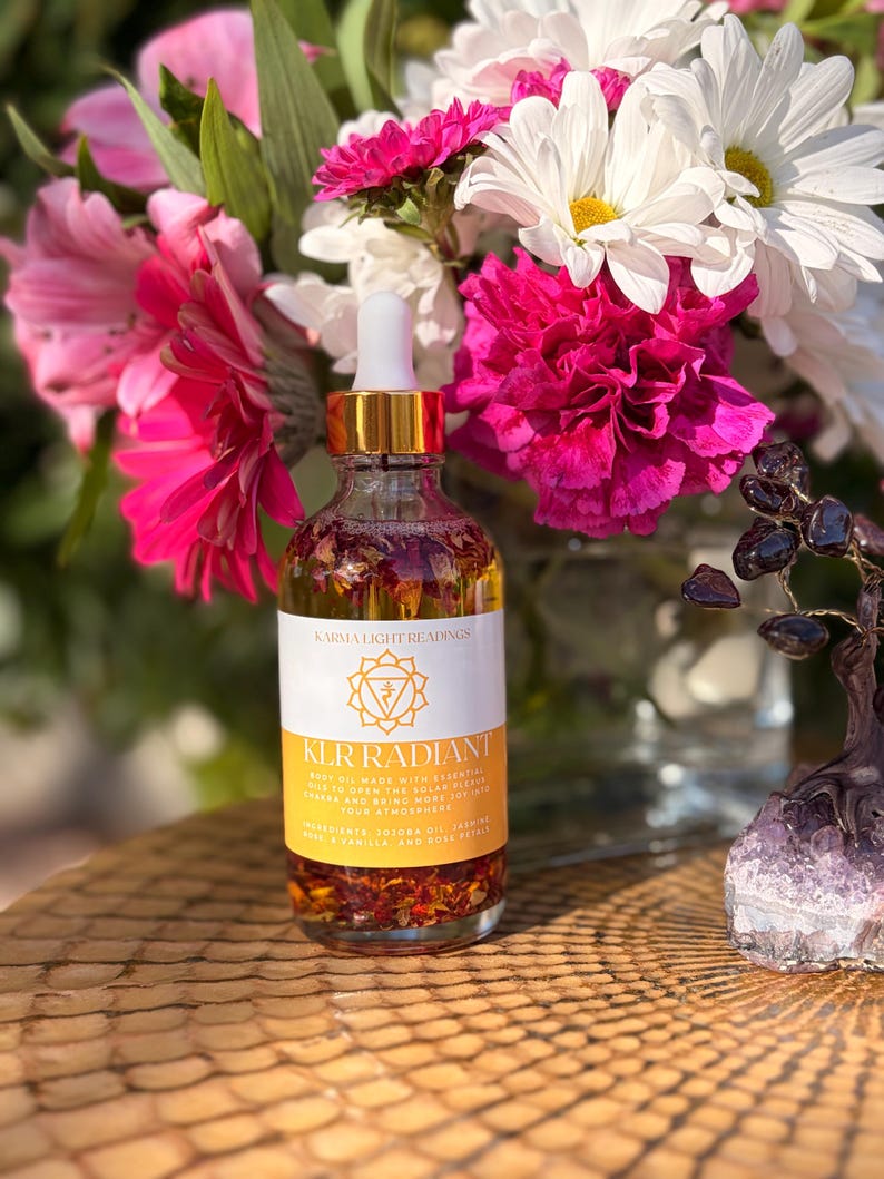 May include: A clear glass bottle with a gold cap and dropper, filled with a golden liquid and flower petals. The label reads "KLR RADIANT" and "Karma Light Readings." The bottle is in front of a bouquet of pink and white flowers.