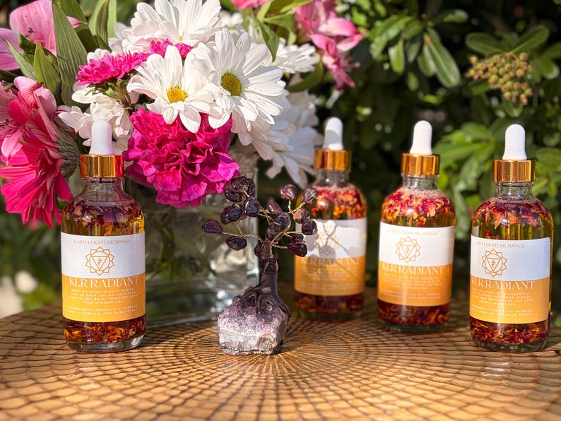 May include: Four glass bottles with gold-colored droppers and labels, filled with a liquid containing flower petals. A small amethyst tree sculpture and a vase of flowers are in the background. The labels read "All Radiant".