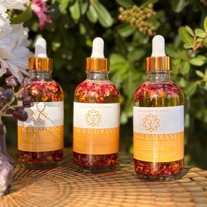 May include: Three glass bottles with gold-colored lids and droppers. The bottles are filled with a clear liquid and flower petals. Each bottle has a white label with the text "KERRADIANT" and a logo. The bottles are on a wooden surface.