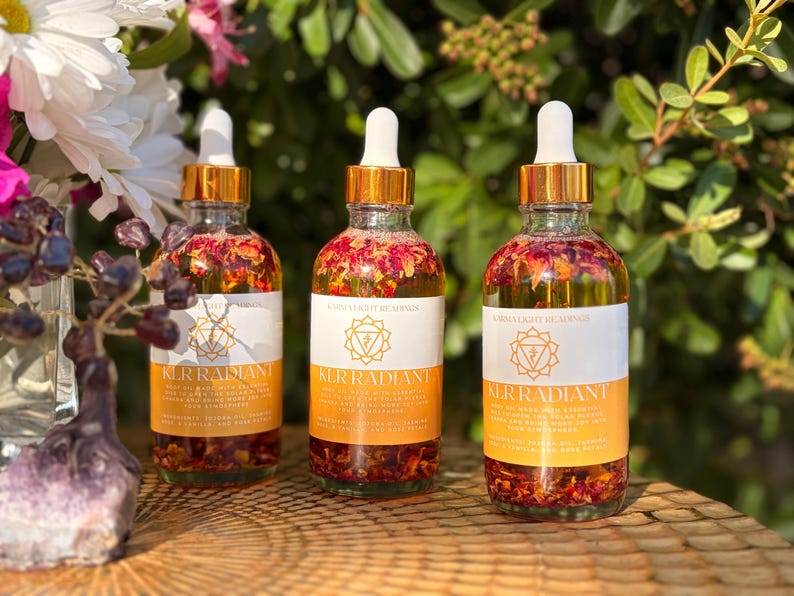 May include: Three glass bottles with gold-colored lids and droppers. The bottles are filled with a clear liquid and flower petals. Each bottle has a white label with the text "KERRADIANT" and a logo. The bottles are on a wooden surface.
