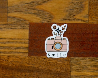 May include: A pink camera with a floral arrangement on top. The camera has a black lens and a white outline. The text "smile" is written below the camera.