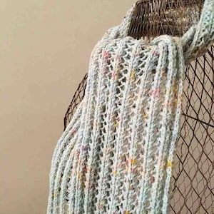 May include: A light blue, open-weave blanket with flecks of pink, yellow, and other colors draped over a wire chair. The blanket has a textured appearance and a soft, inviting look. The chair has a rustic, vintage aesthetic.