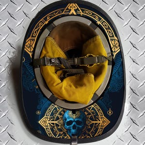 Op de afbeelding: Een brandweerhelm met een blauw en goud decoratief ontwerp. De helm heeft een blauw schedel embleem en ingewikkelde patronen. De binnenkant bevat een gele voering en riemen, met een zilveren metalen sluiting.
