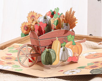 May include: A 3D pop-up card featuring a wheelbarrow filled with pumpkins, gourds, and sunflowers. The card is open and the wheelbarrow is in focus. The background is a light brown surface with a pattern of pumpkins and sunflowers.