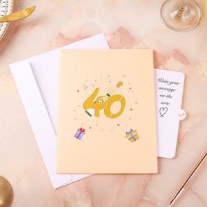 May include: A peach-colored card with a pop-up design featuring the number 40 with a champagne bottle popping out of the zero. The card has confetti and musical notes around the number. The card has a separate note attached with the text "Write your message on the note".
