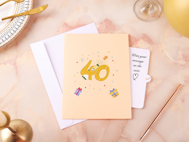 May include: A peach-colored card with a pop-up design featuring the number 40 with a champagne bottle popping out of the zero. The card has confetti and musical notes around the number. The card has a separate note attached with the text "Write your message on the note".