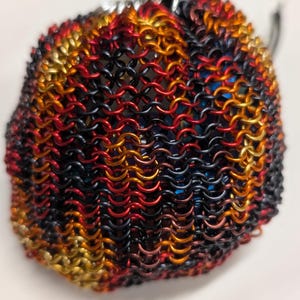 Puede incluir: Una pequeña bolsa redonda con cordón, construida con anillos de metal entrelazados en un patrón de cota de malla. Los anillos presentan un degradado de colores negro, rojo oscuro, naranja y dorado, creando una superficie metálica texturizada.