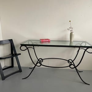 May include: A rectangular glass-topped table with a black wrought iron base. The table features a decorative scroll design. A black folding chair is to the left. A small vase with dried flowers and a book are on the table.