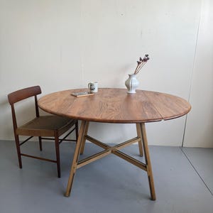 May include: A round wooden dining table with a visible grain pattern and a drop-leaf design, supported by four splayed legs with dark inlay accents. Beside the table sits a wooden chair featuring a woven seat and a curved backrest.