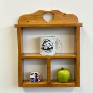 May include: A wooden wall shelf with a heart-shaped cutout, holding a white mug with a black and white illustration, a green apple, and a FISCO Uni-Matic tape measure. The shelf is made of light-coloured wood.