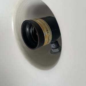 May include: A black light socket with a label, set inside a white, rounded fixture. The label has text that is partially visible. The socket is attached with a nut and bolt.