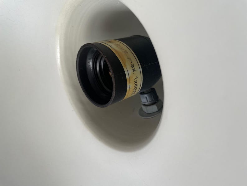 May include: A black light socket with a label, set inside a white, rounded fixture. The label has text that is partially visible. The socket is attached with a nut and bolt.