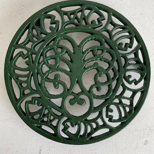 May include: A circular, dark green cast iron trivet with an elaborate, openwork design. The pattern includes a central tree-like motif surrounded by swirling, organic shapes. The trivet is designed to protect surfaces from hot cookware.