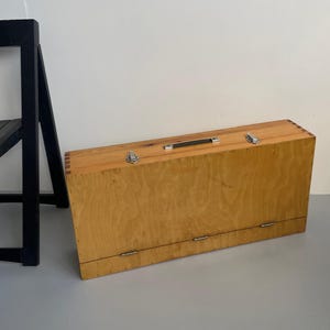 May include: A light-coloured wooden briefcase with a black handle and silver clasps. The rectangular case is closed and has metal hinges along the bottom edge. A black chair leg is visible in the background.