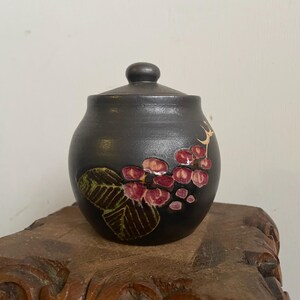 May include: A small, round, black ceramic jar with a lid. The jar is decorated with a floral design in shades of pink, green, and brown.