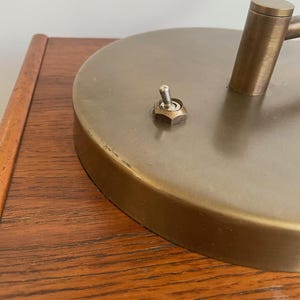 May include: Close-up of a vintage brass desk lamp. The lamp features a circular base with a switch and a cylindrical arm. The lamp is set on a wooden surface.