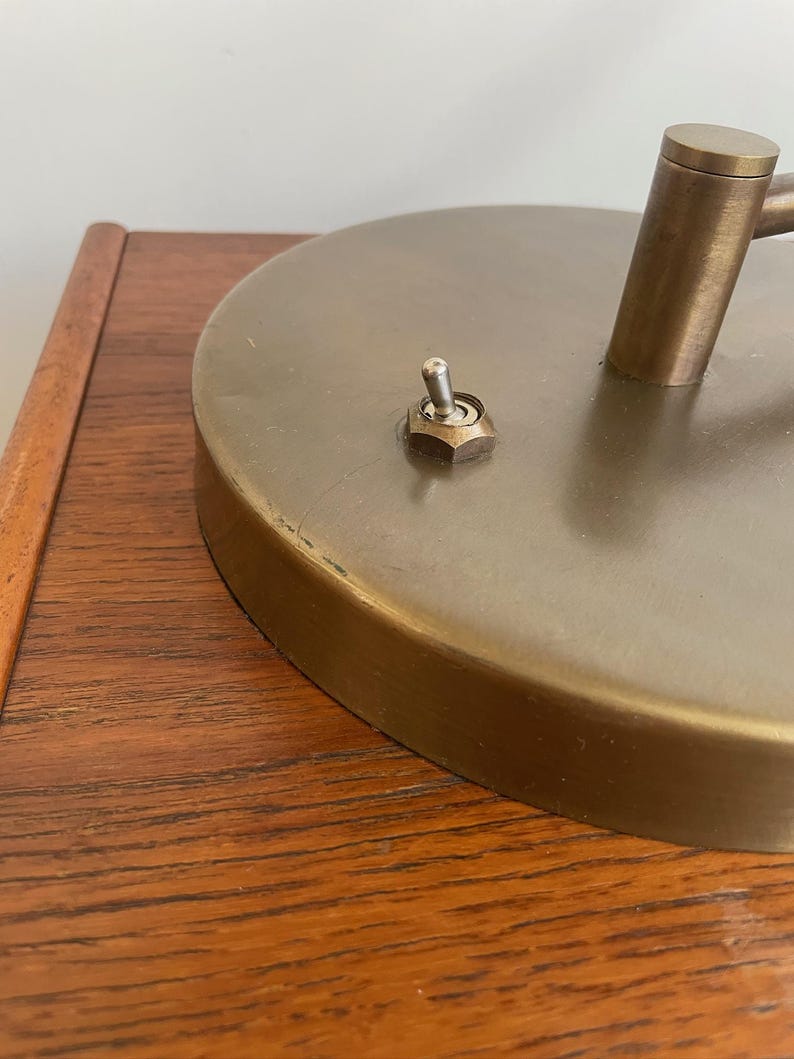 May include: Close-up of a vintage brass desk lamp. The lamp features a circular base with a switch and a cylindrical arm. The lamp is set on a wooden surface.