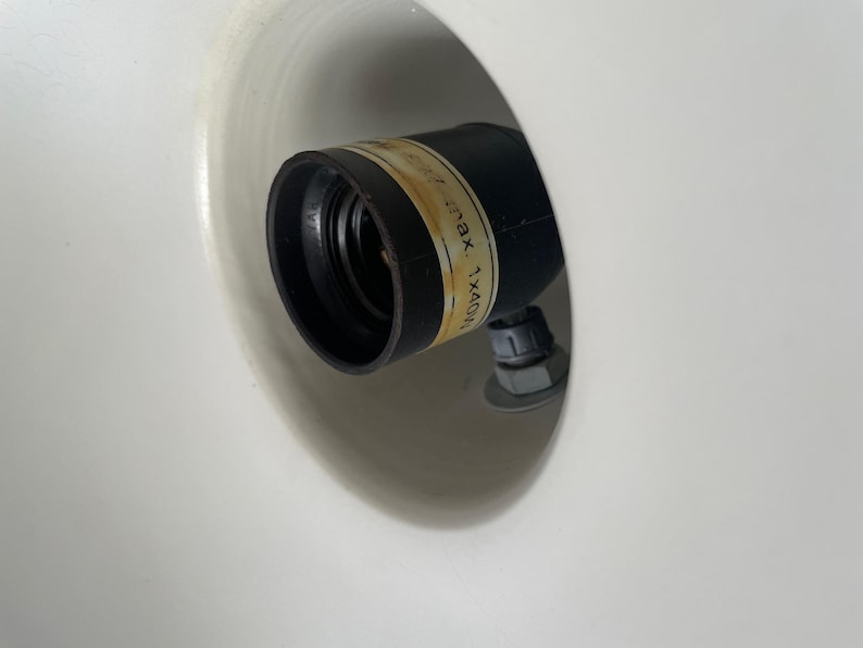 May include: Close-up of a black light socket with a label, set inside a white, rounded fixture. The label has text that is partially visible. The socket is attached with a nut and bolt.