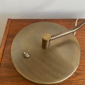 May include: A close-up of a brass-colored desk lamp base with a circular design. The base has a small on/off switch and a curved arm extending upwards. The lamp is resting on a wooden surface.