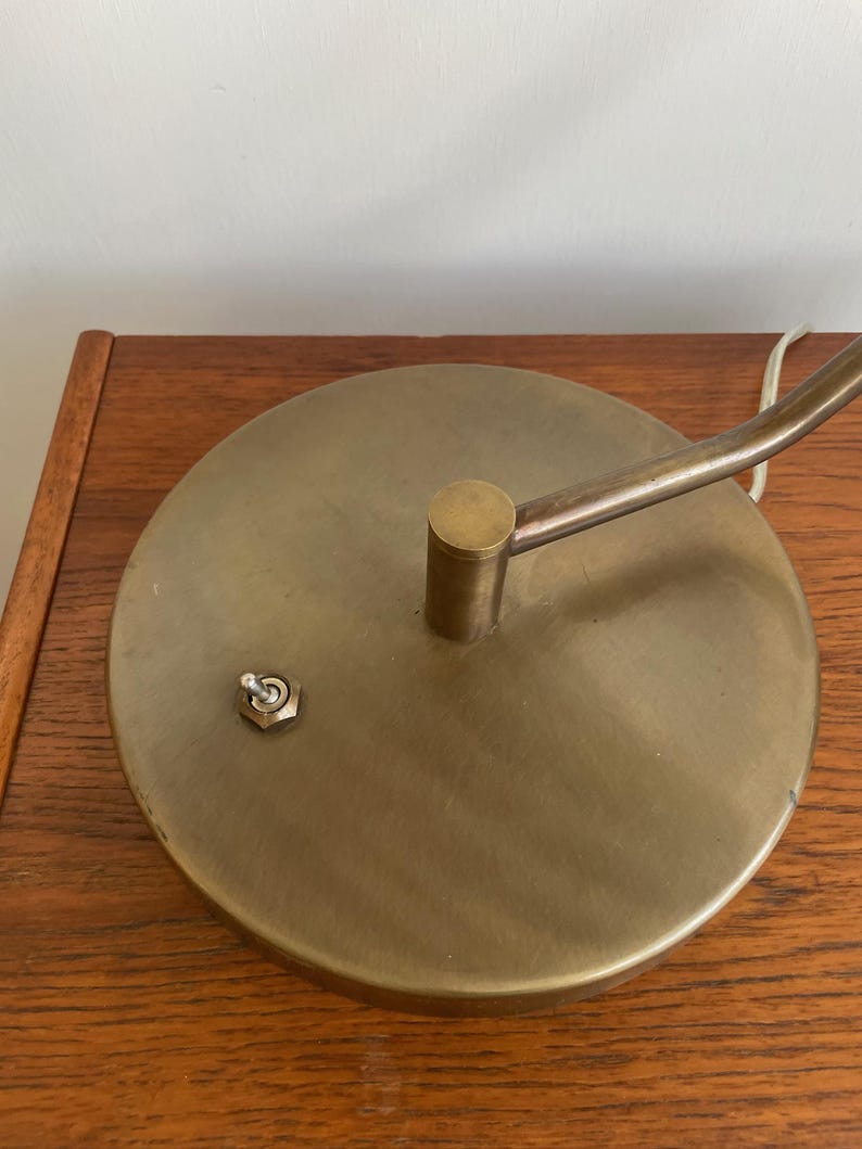 May include: A close-up of a brass-colored desk lamp base with a circular design. The base has a small on/off switch and a curved arm extending upwards. The lamp is resting on a wooden surface.