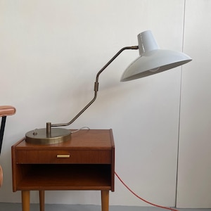 May include: A mid-century modern style bedside table with a drawer and open shelf, made of wood. A white and brass desk lamp with an adjustable arm sits on top. The lamp has a rounded shade and a red power cord.