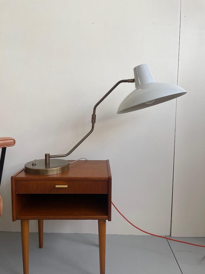 May include: A mid-century modern style bedside table with a drawer and open shelf, made of wood. A white and brass desk lamp with an adjustable arm sits on top. The lamp has a rounded shade and a red power cord.