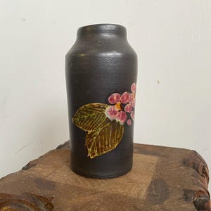 May include: A small black ceramic vase with a floral design. The vase features a pink and yellow flower with green leaves.