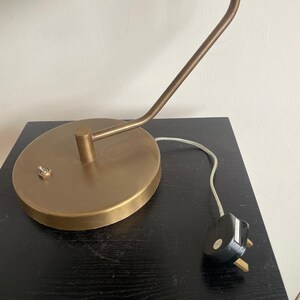 May include: A brass-toned desk lamp with a round base and a curved arm. The lamp has a small switch on the base and a white cord with a black plug. The lamp is on a black surface.