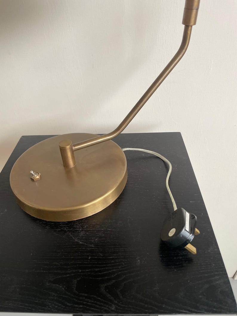 May include: A brass-toned desk lamp with a round base and a curved arm. The lamp has a small switch on the base and a white cord with a black plug. The lamp is on a black surface.