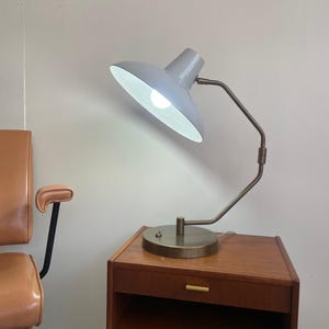 May include: A gray desk lamp with a white interior shade is lit, casting light downward. The lamp has a curved, bronze-colored arm and sits on a wooden nightstand with a drawer. A brown chair is visible in the background.
