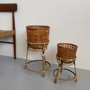 May include: Two woven rattan plant stands. The stands are a natural brown colour and feature a round basket design. The stands have a circular base and are supported by three legs. The larger stand is taller than the smaller one.