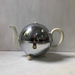 May include: A vintage teapot with a spherical, chrome-plated body and white ceramic spout, handle, and lid. The teapot stands on three small, rounded feet. The chrome surface reflects the surroundings.