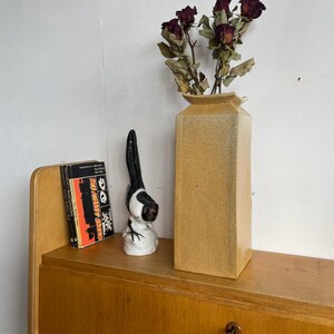 May include: A ceramic vase with a rectangular shape and a light brown glaze. The vase is filled with dried red roses. A small ceramic figurine of a magpie is on a shelf behind the vase.
