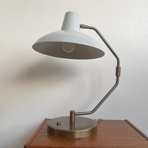 May include: A white and brass desk lamp with a curved arm and a round base. The lamp has a white lampshade and a brass-colored arm and base. The lamp is sitting on a wooden nightstand.