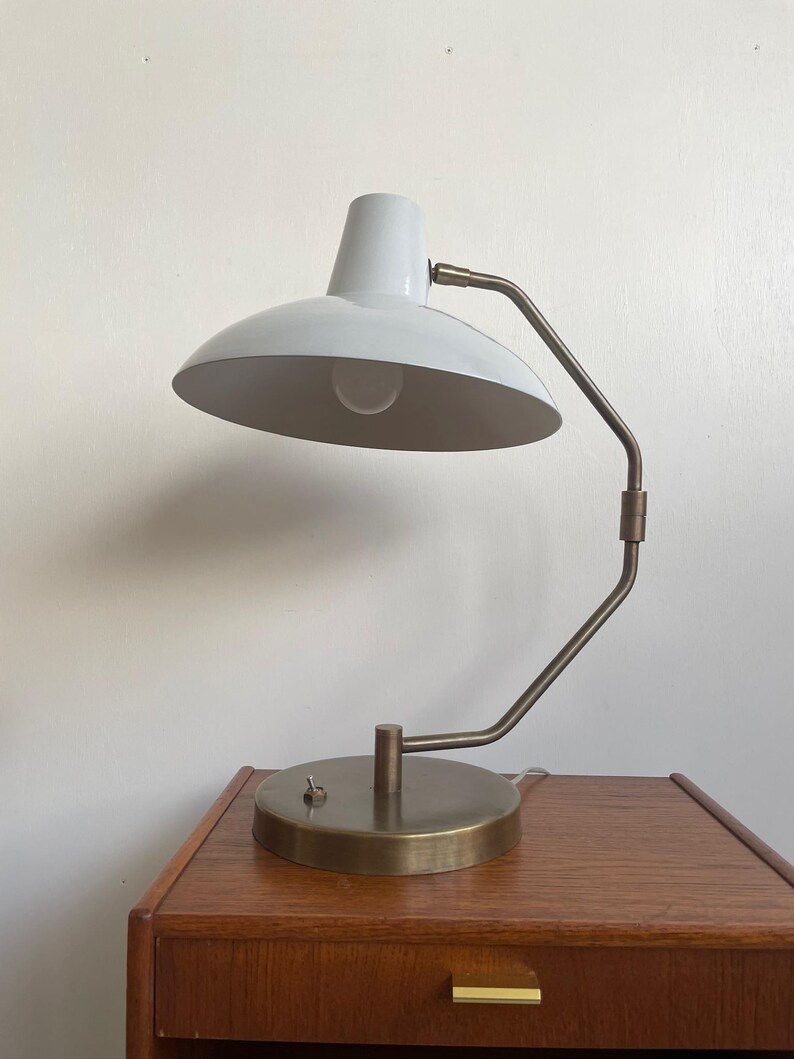 May include: A white and brass desk lamp with a curved arm and a round base. The lamp has a white lampshade and a brass-colored arm and base. The lamp is sitting on a wooden nightstand.