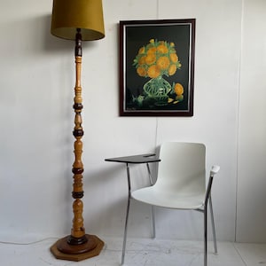 May include: A wooden floor lamp with a green lampshade and a brown wooden base. The lamp is next to a white chair with a black desk attached to the armrest. A framed picture of yellow flowers in a vase is on the wall behind the chair.