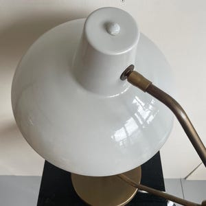 May include: A white desk lamp with a rounded shade and a brass-colored arm and base. The lamp is positioned on a black surface, with a white wall in the background. The lamp's design is modern and minimalist.