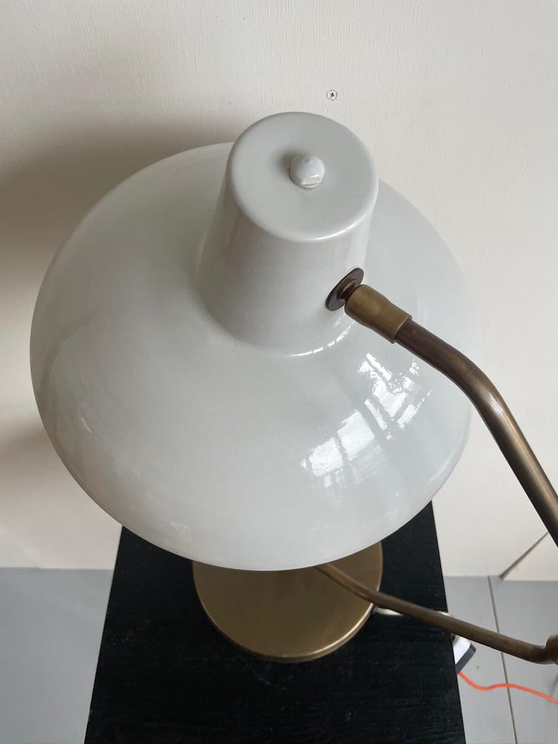 May include: A white desk lamp with a rounded shade and a brass-colored arm and base. The lamp is positioned on a black surface, with a white wall in the background. The lamp's design is modern and minimalist.