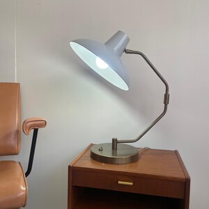 May include: A gray desk lamp with a white interior, positioned on a wooden nightstand. The lamp has a curved arm and a circular base. The nightstand has a drawer with a gold-colored handle and a lower shelf.