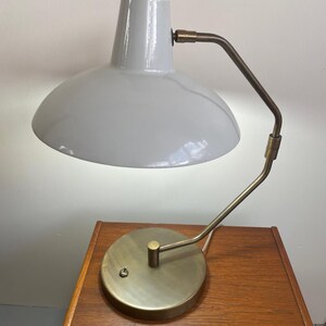 May include: A gray desk lamp with a curved brass arm and a circular base. The lamp has a rounded shade and a small switch on the base. The lamp is sitting on a wooden nightstand.