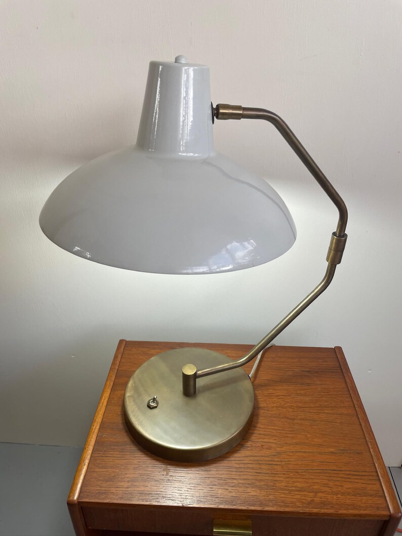 May include: A gray desk lamp with a curved brass arm and a circular base. The lamp has a rounded shade and a small switch on the base. The lamp is sitting on a wooden nightstand.