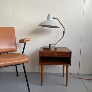 May include: A mid-century modern nightstand with a white lamp and a brown leather chair. The nightstand is made of wood with a drawer and a lower shelf. The lamp has a white shade and a brass-colored arm.