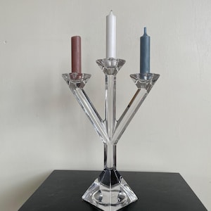 May include: A clear crystal candelabra with three candle holders. It holds a brown, white, and blue candle. The base is a geometric shape, and the arms extend upwards to hold the candles. The candelabra is on a black surface.