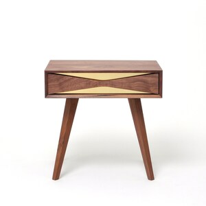 May include: A walnut wood nightstand with a brass drawer front. The drawer has a unique curved design.