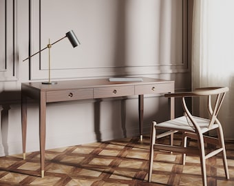 May include: A wooden desk with three drawers and brass hardware. The desk has a black lamp with a gold base and a laptop on top. A wooden chair with a woven seat is positioned next to the desk. The desk and chair are in a room with a light pink wall and a patterned floor.