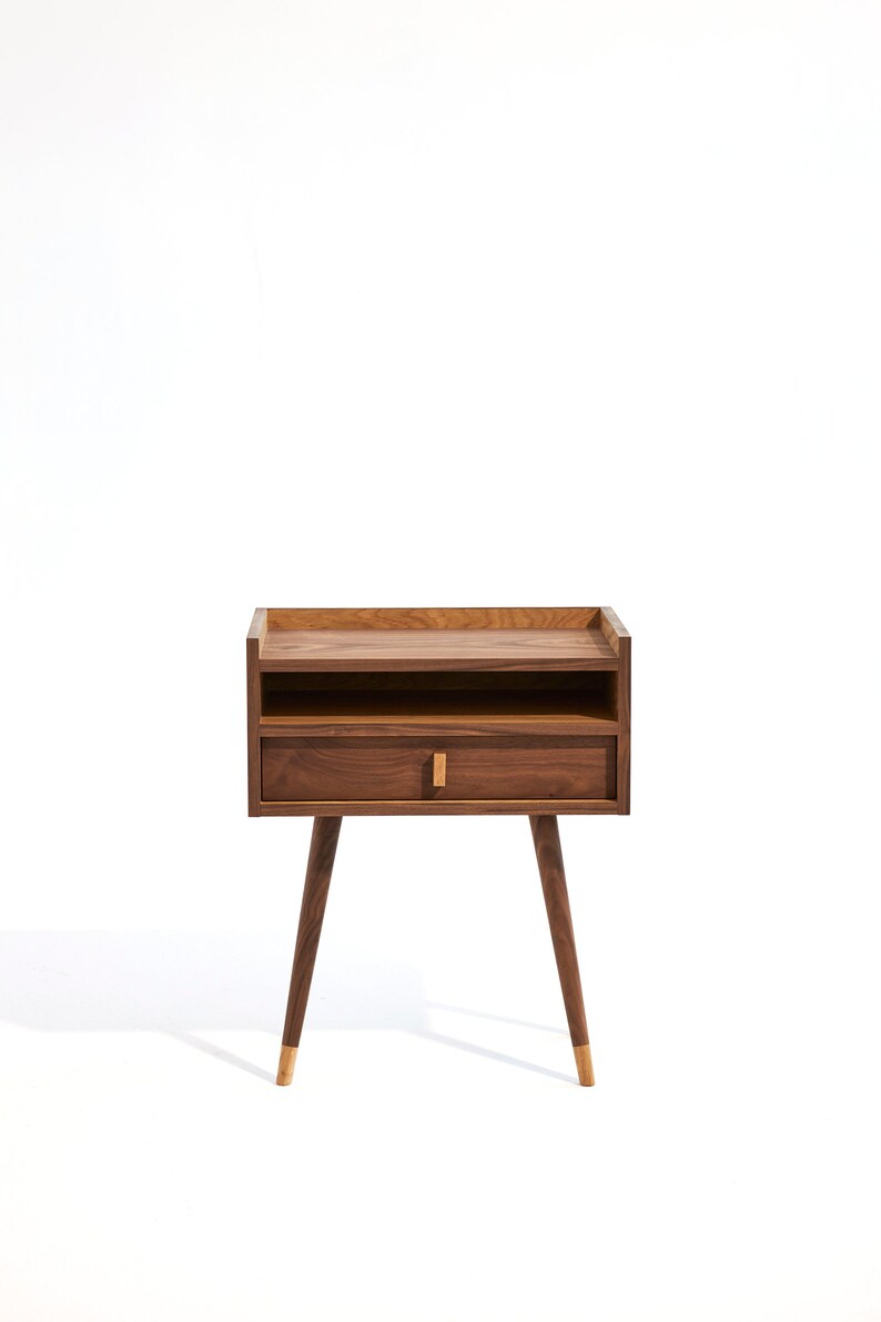 May include: A wooden nightstand with a single drawer and a shelf. The nightstand has a dark brown finish and gold-colored legs.