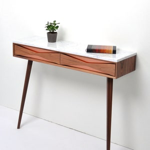 May include: A contemporary wooden console table with a white marble top and two drawers. The drawers feature a distinctive wave design. The table has four dark brown, angled legs. A small potted plant and a book are on the table.
