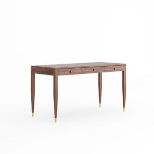 May include: A rectangular dark brown wooden desk featuring three horizontal drawers with small circular knobs. The desk is supported by four tapered legs, each finished with a metallic gold-toned cap at the bottom, providing a clean and structured appearance.
