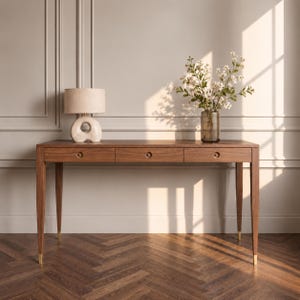 May include: A rectangular wooden desk with three drawers, each featuring a circular pull. The desk has four tapered legs with gold-toned metal caps at the feet. A beige lamp with a rounded base and a glass vase with white flowers sit on top.