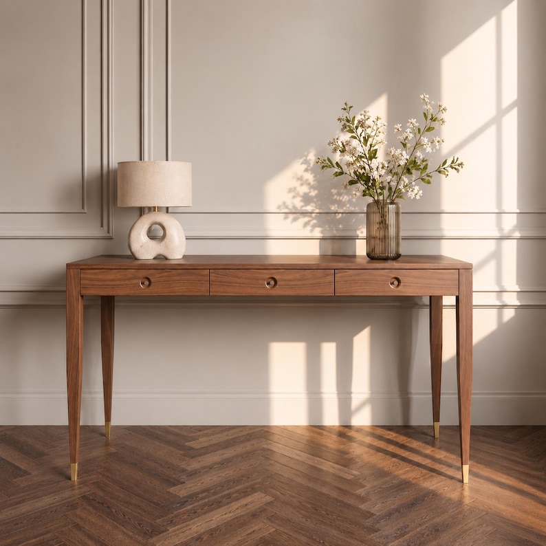May include: A rectangular wooden desk with three drawers, each featuring a circular pull. The desk has four tapered legs with gold-toned metal caps at the feet. A beige lamp with a rounded base and a glass vase with white flowers sit on top.